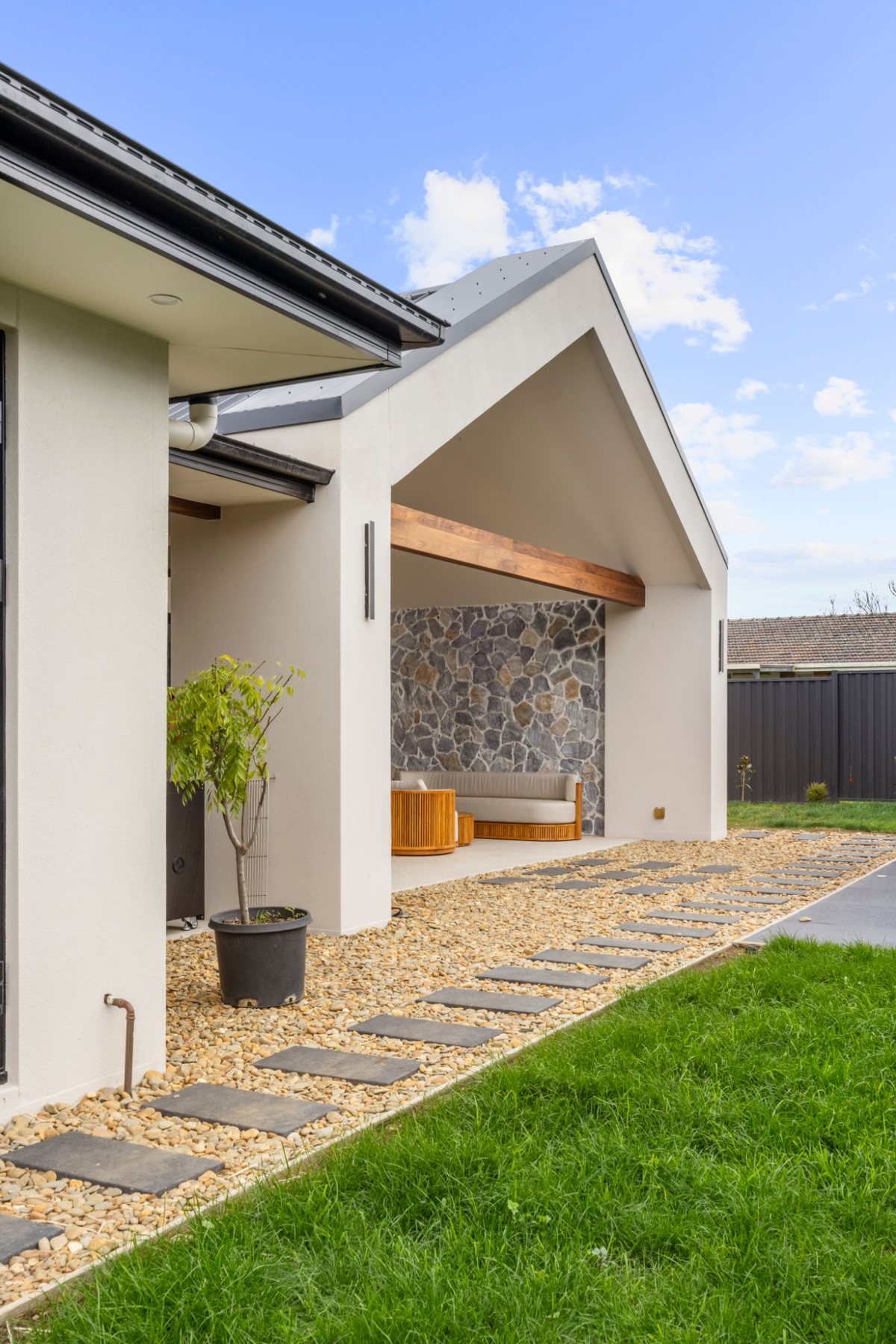Gabled outdoor room with a timber beam and stacked stone feature wall, beside a stepping-stone path through a pebble garden