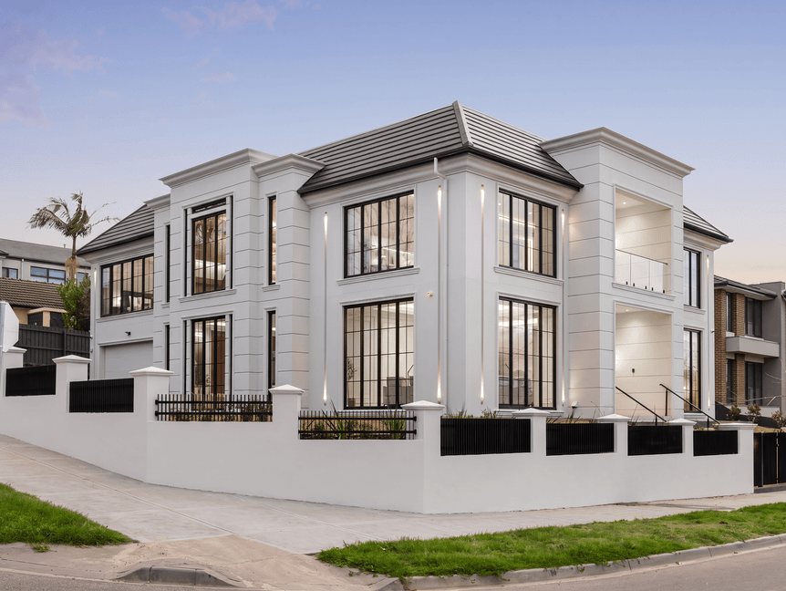 Two-storey rendered home with classical detailing and tall black-framed windows, behind a rendered front fence on a corner block
